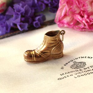 May include: A small, gold-colored boot charm with intricate detailing. The charm has a loop at the top for attachment and is set against a soft, textured background. The boot's design includes laces and a sturdy sole, suggesting a miniature work boot.
