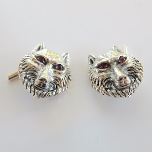 May include: A pair of silver-toned cufflinks, each shaped like a wolf's head. The wolf's eyes are small, red gemstones. The cufflinks are detailed with fur texture and have a classic post and toggle closure.