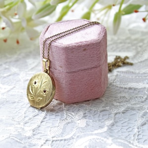 May include: A gold-toned circular locket pendant on a delicate matching chain. The locket features a brushed metal finish with an engraved floral motif and two small, embedded red stones at the centre of the flower petals, resting against a pink box.