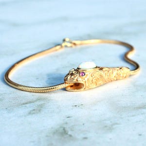 以下が含まれることがあります： 金色の蛇のブレスレット。装飾的な頭のデザイン。蛇の頭には楕円形の白い石と小さなピンク色の宝石が付いています。ブレスレットは柔軟で質感のあるチェーンです。
