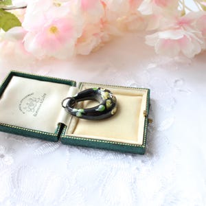 May include: A black glass ring with hand-painted floral details in shades of green, white, and yellow. The ring is displayed in a green velvet box with a white satin lining.