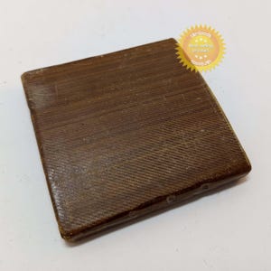 Puede incluir: Un objeto rectangular marrón texturizado con un patrón tejido. El objeto tiene una apariencia ligeramente desgastada y está sobre un fondo blanco. Una pegatina amarilla con texto es visible en la esquina superior derecha.