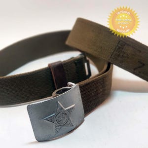 Peut inclure: Une ceinture en toile vert olive dotée d'une boucle en métal argenté avec un emblème en forme d'étoile à cinq branches en relief. La ceinture comprend un passant en cuir marron foncé et un embout métallique à l'extrémité de la sangle.