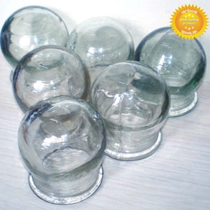 Op de afbeelding: Een verzameling van zes heldere glazen cupping therapie cups. Elke cup is bolvormig met een platte, cirkelvormige basis. De cups zijn in een groep gerangschikt, wat hun transparantie en eenvoudige ontwerp benadrukt. De afbeelding bevat een sticker met de tekst "UKR Goods Best-selling product".