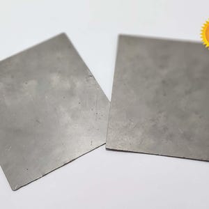 May include: Two rectangular, gray metal plates are displayed against a white background. The plates have a slightly textured surface and appear to be made of a solid, durable material. A yellow sticker with text is visible in the upper right corner.