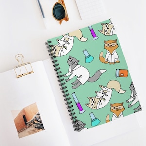 May include: A green spiral notebook featuring a repeating pattern of cartoon cats dressed in lab coats and holding beakers and test tubes. The cats are depicted in various poses, some standing, some sitting, and some lying down. The background is a light green colour.