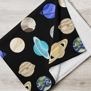 Puede incluir: Una manta negra con un patrón repetido de planetas coloridos, incluyendo Saturno, Júpiter y la Tierra. Los planetas están representados en tonos de azul, marrón y beige. La manta tiene una parte inferior blanca.