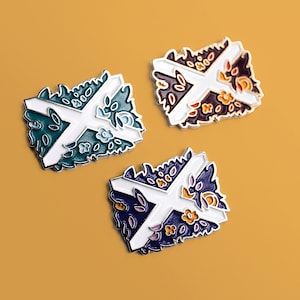 以下が含まれることがあります： 白い十字架に花柄のデザインが施されたエナメルピンが3つ。ピンはターコイズ、パープル、オレンジです。