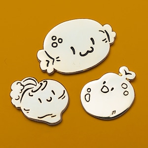 May include: Three white enamel pins with black outlines depicting cartoon faces. The pins are shaped like a round bun, a heart, and a teardrop.
