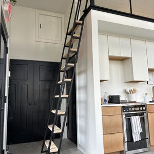 May include: A black metal ladder with wooden steps leading up to a loft. The ladder is in a room with white walls and a black door. There are wooden cabinets and a stainless steel oven in the background.