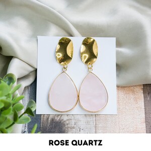 Op de afbeelding: Een paar gouden oorbellen met roze kwarts. De oorbellen hebben een traanvormige roze kwarts steen met een gouden zetting. De oorbellen zijn op een witte achtergrond.