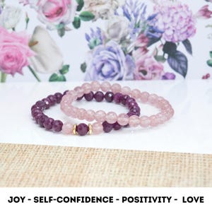 May include: Two bracelets, one pink and one purple, with gold accents. The bracelets are made of small beads and are stacked on top of each other. The text "JOY - SELF-CONFIDENCE - POSITIVITY - LOVE" is printed below the bracelets.