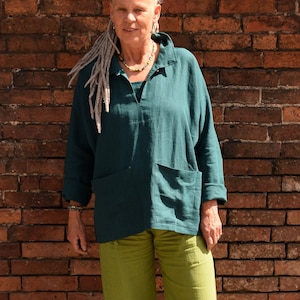 May include: A woman wearing a teal linen shirt with a  patch pocket and matching green linen pants. She is standing in front of a brick wall and a small crocheted stool.