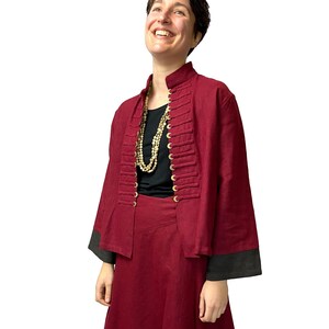 Op de afbeelding: Een vrouw draagt een bordeauxrode rok en jas met gouden knopen. Ze draagt een zwart topje met een gouden ketting en bordeauxrode laarzen.