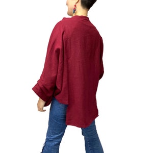 May include: A burgundy-colored, long-sleeved top with a relaxed fit. The top features a wide, open design and a slightly asymmetrical hemline. The sleeves are wide and gathered at the cuffs. The model is wearing blue jeans.