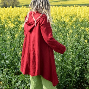 Pode incluir: Uma jaqueta de linho vermelha com capuz e um design longo e fluido. A jaqueta é usada por uma pessoa com longos dreadlocks cinzentos em pé num campo de flores amarelas.