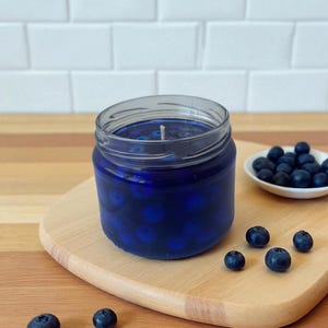 May include: A blueberry-scented candle in a clear glass jar, filled with blue wax and whole blueberries. The candle sits on a wooden cutting board, with fresh blueberries scattered around and in a small white dish.