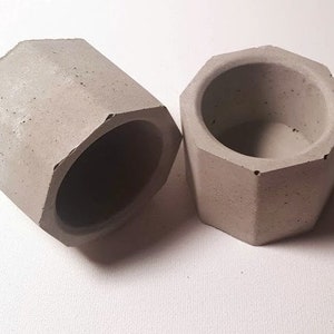 May include: Two gray concrete hexagonal planters. The planters are made of a rough, textured material and have a slightly rounded lip.