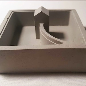 May include: A gray concrete planter with a geometric design. The planter has a small house-shaped structure and a curved pathway inside.