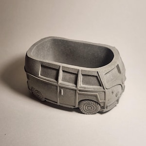 May include: A gray concrete planter shaped like a vintage van. The planter has detailed features including windows, wheels, and a front grill. The open top provides space for small plants or succulents. The overall design is a retro, minimalist style.