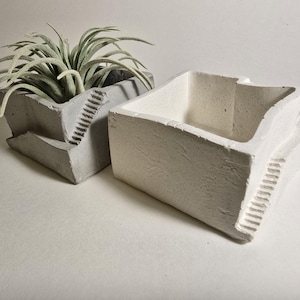 May include: Two concrete planters with a geometric design. The planter on the left is gray and has a plant inside. The planter on the right is white.