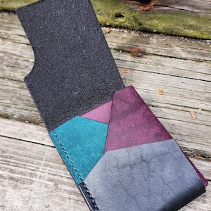May include: Handmade leather wallet with a geometric design. The wallet features a black exterior and interior, with teal, purple, and gray color-blocked sections. The wallet is open, revealing the interior pockets, and is set against a wooden background.