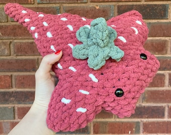 May include: A handmade, plush strawberry pillow in shades of red and green. The pillow features white speckles, a green leaf cluster, and black button eyes. The pillow is approximately 12 inches long.