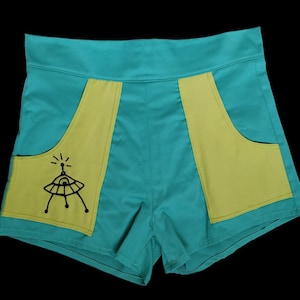 Puede incluir: Pantalones cortos turquesa de cintura alta con detalles de bolsillo verde lima. Un bolsillo presenta un gráfico negro de un OVNI. Los pantalones cortos están hechos de una tela suave y tienen una cintura ancha. El estilo general es retro y juguetón.