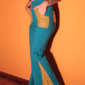 Puede incluir: Pantalones acampanados turquesa y dorados con detalles adornados. Los pantalones de cintura alta presentan detalles dorados en los bolsillos y paneles acampanados en las piernas. Los pantalones se combinan con un top corto color cobre. El fondo es naranja liso.