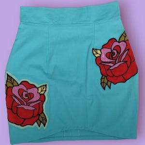 Può includere: Minigonna turchese con due grandi rose rosse ricamate con centri rosa e foglie verdi e dorate. La gonna ha una vita alta e un design semplice e su misura.