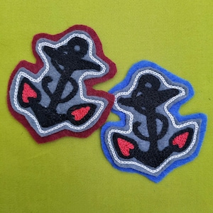 Puede incluir: Dos parches de fieltro con anclas bordadas. Un parche es rojo con un contorno blanco y el otro es azul con un contorno blanco. Ambas anclas son negras con corazones rojos en la parte inferior.