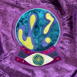 Op de afbeelding: Een geborduurd embleem in paars, turquoise en geel met een kristallen bol. De kristallen bol heeft een turquoise achtergrond met gele en blauwe vormen. De basis van het embleem heeft een paarse band met rode accenten en een oogontwerp.
