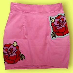 Op de afbeelding: Een roze minirok met twee geborduurde rozenapplicaties. De rozen zijn rood met roze centra, omlijnd in zwart, en hebben groene bladeren. De rok is gemaakt van een gladde, glanzende stof.