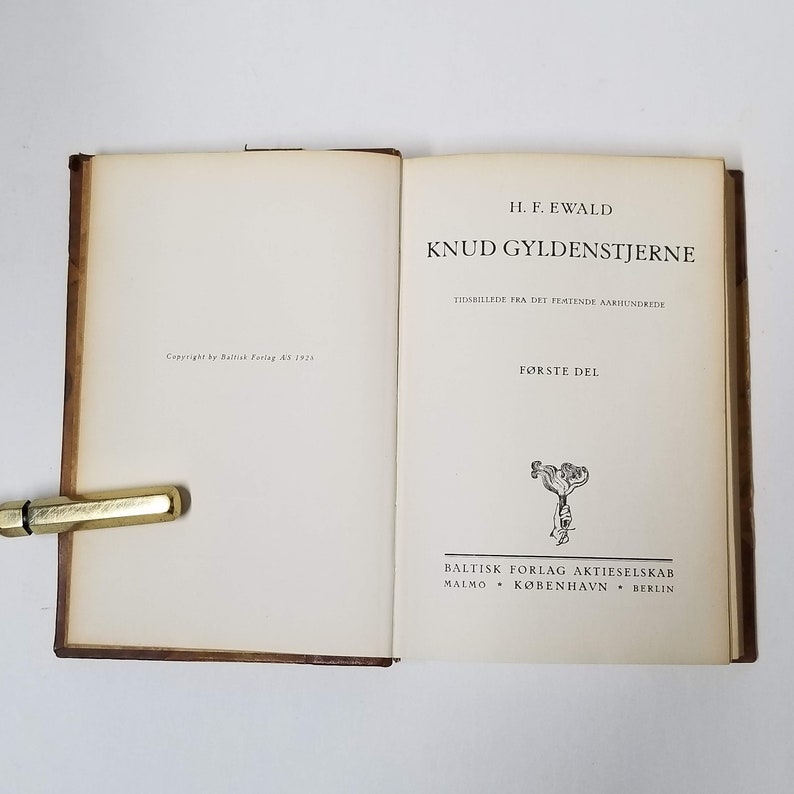 Puede incluir: Un libro abierto con el t&iacute;tulo "KNUD GYLDENSTJERNE" de H.F. Ewald. Las p&aacute;ginas son blanquecinas, con texto negro. La cubierta es marr&oacute;n. Un bol&iacute;grafo dorado descansa sobre la p&aacute;gina izquierda. Publicado por Baltisk Forlag Aktieselskab en 1925.