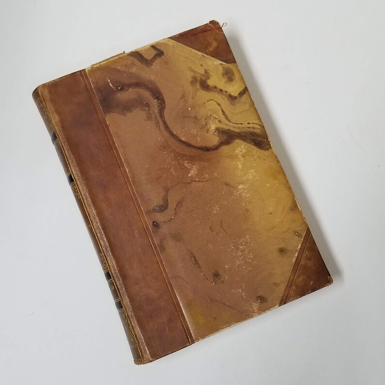 Puede incluir: Un libro antiguo con una cubierta veteada en tonos marrones y beige. El lomo y las esquinas son de un marr&oacute;n m&aacute;s oscuro. El libro muestra signos de desgaste, lo que sugiere que es un art&iacute;culo antiguo. Los bordes de las p&aacute;ginas son visibles.