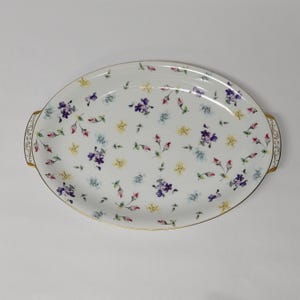 May include: Oval serving platter with a white background and a floral pattern of purple, yellow, and blue flowers. The platter has gold trim and decorative handles. A delicate and elegant piece for serving food.