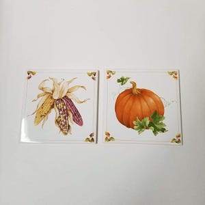 May include: Two square ceramic tiles with autumn-themed illustrations. One tile features two ears of corn, one yellow and one purple, with tan husks. The other tile shows a large orange pumpkin with green leaves and vines.