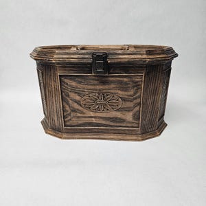 May include: A wooden chest with a dark brown finish and a decorative floral carving on the front panel. The chest has a hinged lid and a metal clasp. The chest is octagonal in shape and has a handle on top.