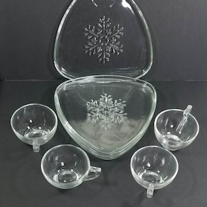 May include: A clear glass serving set with a snowflake design. The set includes a triangular serving platter, a triangular bowl, and four small cups with handles. The snowflake design is embossed on the platter and bowl.