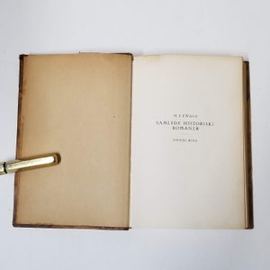 Puede incluir: Un libro abierto con p&aacute;ginas envejecidas de color blanquecino y una cubierta marr&oacute;n. La p&aacute;gina del t&iacute;tulo presenta el texto "H.F. EWALD SAMLEDE HISTORISKE ROMANER TIENDE BIND" en negro. Un bol&iacute;grafo dorado descansa en el borde del libro.
