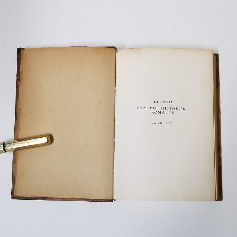 Puede incluir: Un libro abierto con p&aacute;ginas envejecidas de color blanquecino y una cubierta marr&oacute;n. La p&aacute;gina del t&iacute;tulo presenta el texto "H.F. EWALD SAMLEDE HISTORISKE ROMANER TIENDE BIND" en negro. Un bol&iacute;grafo dorado descansa en el borde del libro.
