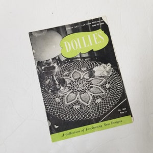 May include: Vintage "DOLLIES" booklet with a black and green cover, featuring a white crocheted doily. The cover text includes "CLARK'S O.N.T. J&P COATS BOOK No. 235" and "A Collection of Fascinating New Designs". A lamp is visible in the background.