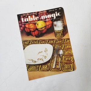 May include: A vintage magazine cover titled "Table Magic" featuring a still life of fruit in a wire basket, a glass of liquid, and a place setting with a plate, knife, and spoon. The magazine cover includes the text "PLACE MATS GLASS JACKETS BOTTLE CAP CROCHET".
