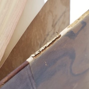Puede incluir: Primer plano de un libro antiguo con una cubierta marr&oacute;n desgastada y p&aacute;ginas envejecidas. Los bordes de la cubierta muestran signos de desgaste, con pintura descascarillada y madera expuesta. Las p&aacute;ginas son de color marr&oacute;n claro.
