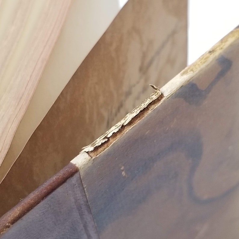 Puede incluir: Primer plano de un libro antiguo con una cubierta marr&oacute;n desgastada y p&aacute;ginas envejecidas. Los bordes de la cubierta muestran signos de desgaste, con pintura descascarillada y madera expuesta. Las p&aacute;ginas son de color marr&oacute;n claro.