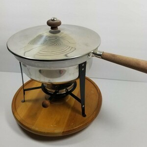 May include: A vintage fondue pot with a stainless steel pot and lid, a wooden handle, and a wooden knob. The pot sits on a wooden base with a black metal stand. The fondue pot is designed for cooking and serving.