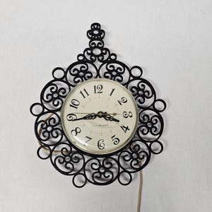 May include: A wall clock featuring a circular off-white face with black Arabic numerals and ornate black metal scrollwork surrounding the dial. The clock includes black hands and a General Electric logo on the face, with a power cord extending downward.