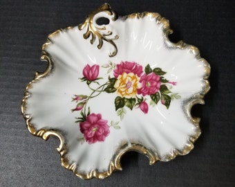 May include: White porcelain dish with a ruffled edge and gold trim. The dish features a floral design with pink and yellow roses and green leaves. A gold handle is on the side.