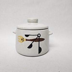May include: A white ceramic lidded jar with a speckled finish and two small side handles. The front features a graphic illustration of a yellow fork and three black measuring spoons arranged in a cluster, set against the off-white speckled surface.