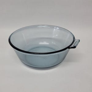May include: A clear, round, glass bowl with a light blue tint. The bowl has a black rim and a handle on one side. The bowl is empty and sits on a white surface. The bowl is likely for food preparation or serving.